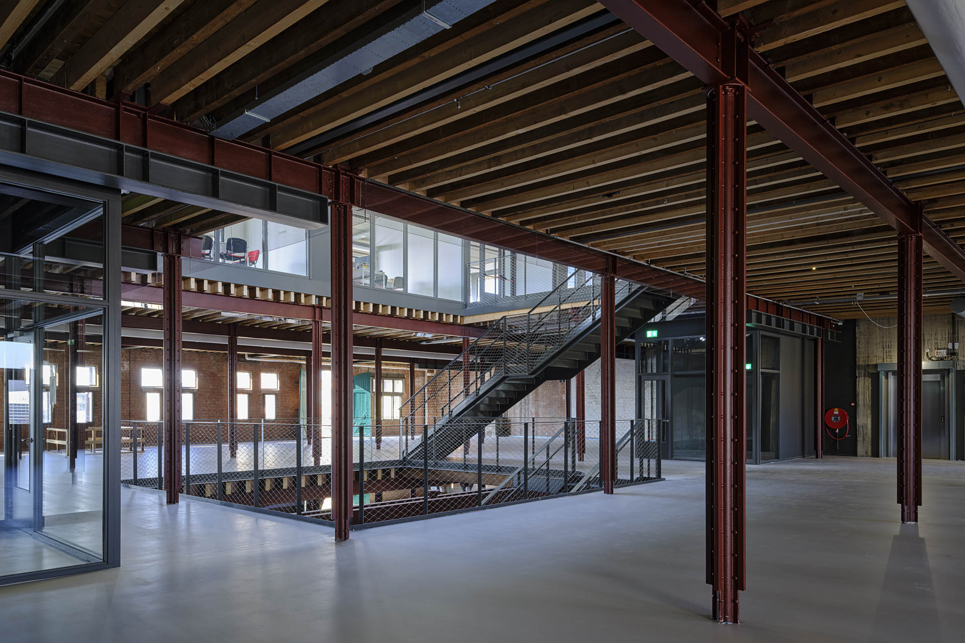 Ein industrieller Innenraum mit bordeauxfarbenen Stahlstützen und betonierten Decken. In der Mitte ein Atrium, das zu den oberen und unteren Geschossen führt. Eine Stahltreppe verbindet die Ebenen, und die Wände sind aus Ziegeln.
