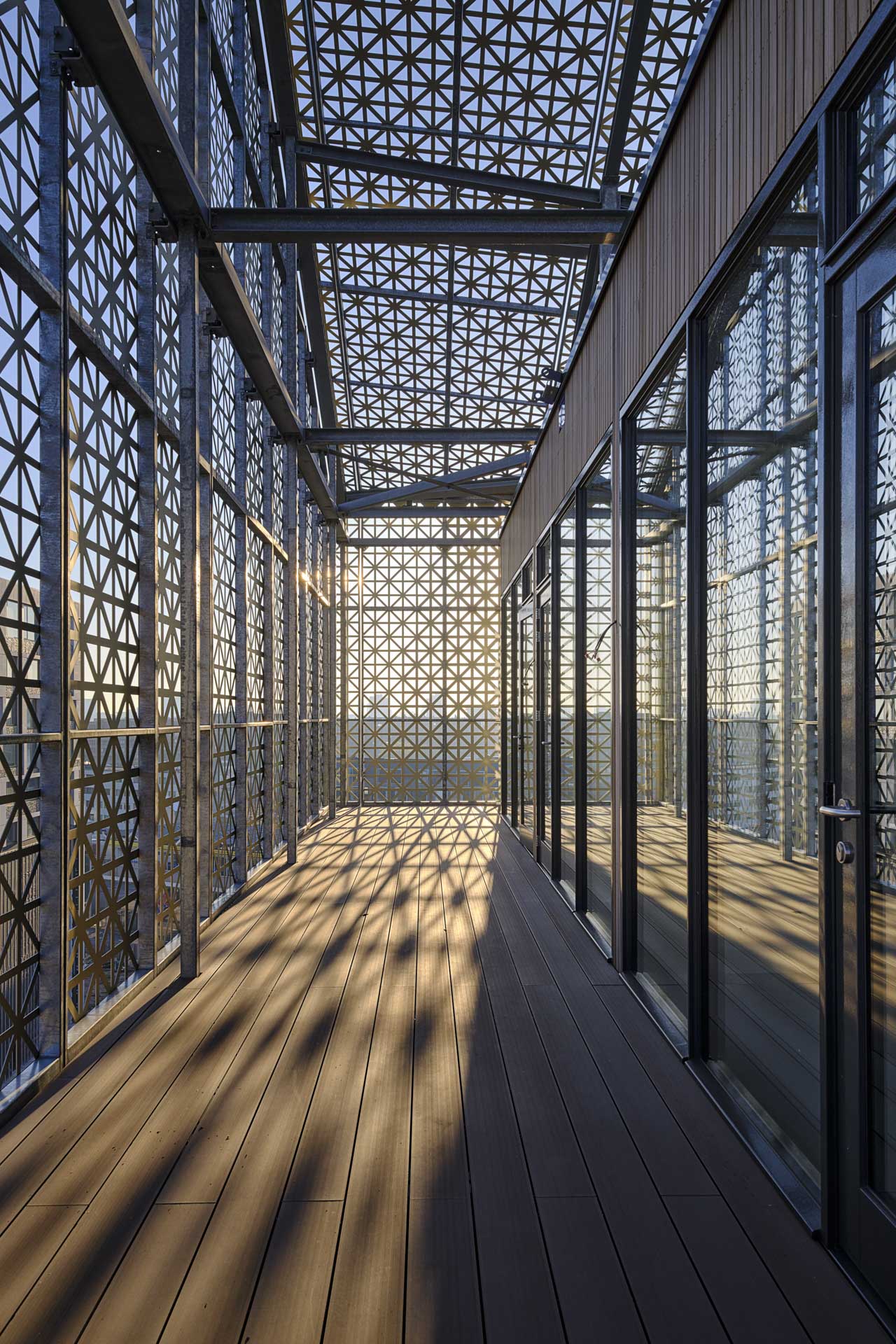 Der Dachaufbau aus Aluminium-Lochpaneelen erzeugt ein Lichtspiel aus Sonnenmustern auf den umlaufenden Laubengängen mit Holzboden. Die Holzfassade ist mit vielen Terrassentüren versehen, die auf die Laubengangbalkone führen.