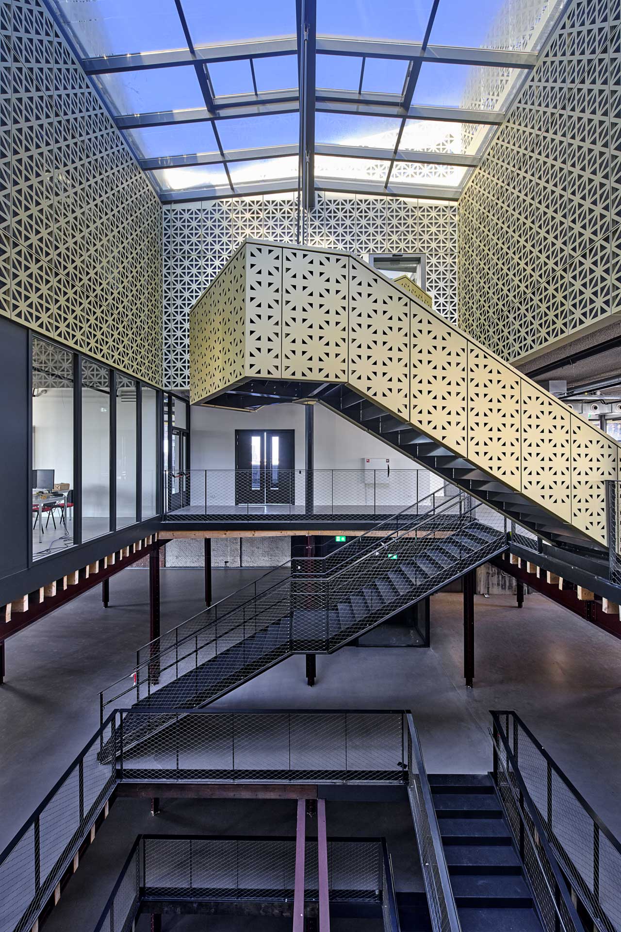 Der Innenraum zeigt einen Deckendurchbruch über alle Ebenen, darüber spannt sich ein Glasdach. Stahltreppen verbinden die Geschosse. Das oberste Geschoss ist mit gemusterten Metallpaneelen verziert und hebt sich von den industriellen Elementen ab.