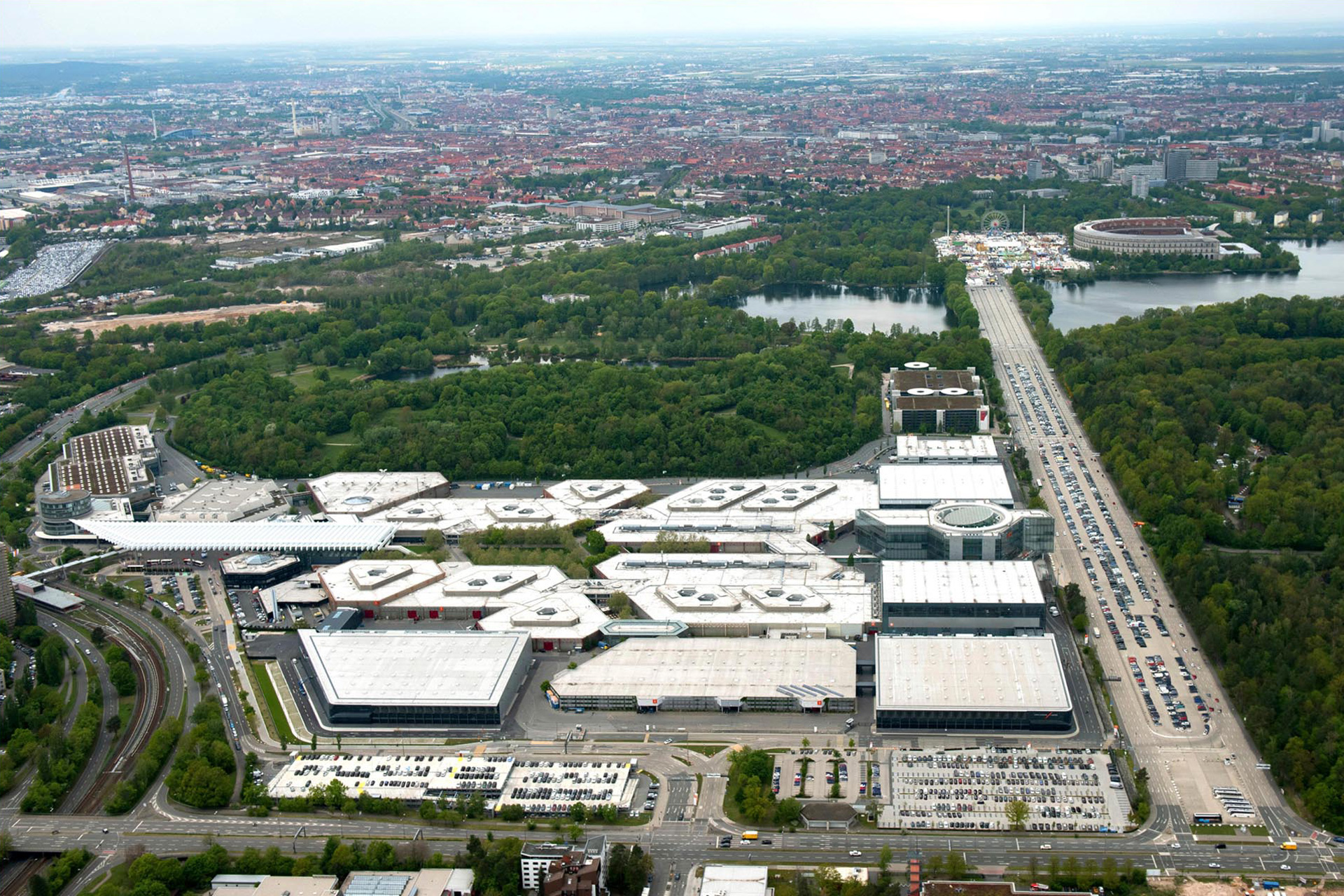 Luftaufnahme des Messe N&uuml;rnberg mit seinen diversen Hallen. Rechts erkennt man einen gro&szlig;en Parkplatz, dahinter ein kleines Waldst&uuml;ck bevor die Stadt zu sehen ist.