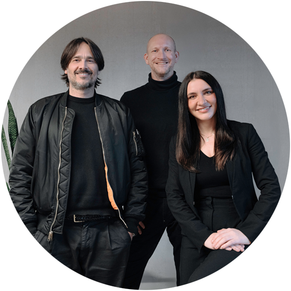 Zwei M&auml;nner und eine Frau l&auml;cheln in die Kamera. Alle sind in schwarz gekleidet und stehen vor einem grauen Hintergrund.